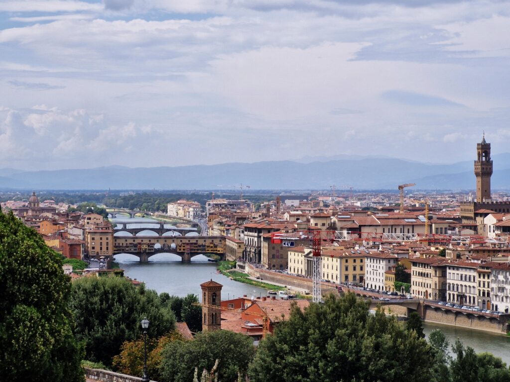 Toskania co warto zobaczyć. Panorama Florencji z widokiem na rzekę Arno, słynny most Ponte Vecchio oraz wieżę Palazzo Vecchio, otoczona wzgórzami Toskanii pod zachmurzonym niebem.