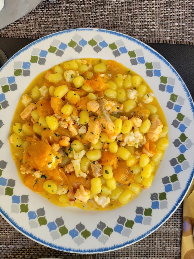 : A top-down view of a patterned plate filled with a yellow-tinted Italian pasta dish, featuring small, round cavatelli-style pasta, chickpeas, pieces of squid, salmon, and yellow tomatoes, garnished with herbs.