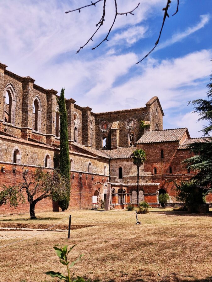 A courtyard view of the medieval Abbey of San Galgano ruins in Tuscany, featuring red brick lower walls, towering stone walls with arched gothic windows, a tall cypress tree, and a small palm tree under a bright blue sky.