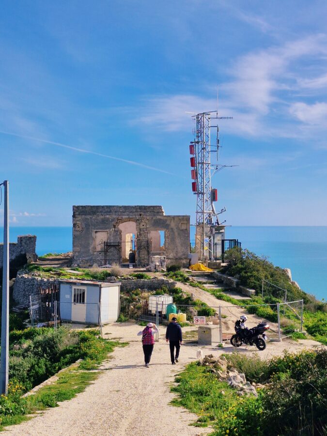 Two people and a small white dog walk down a dirt path toward the stone ruins of an old building on a coastal cliff. Next to the structure stands a tall metal telecommunications tower with red antennas, with a clear blue sea and sky in the background.