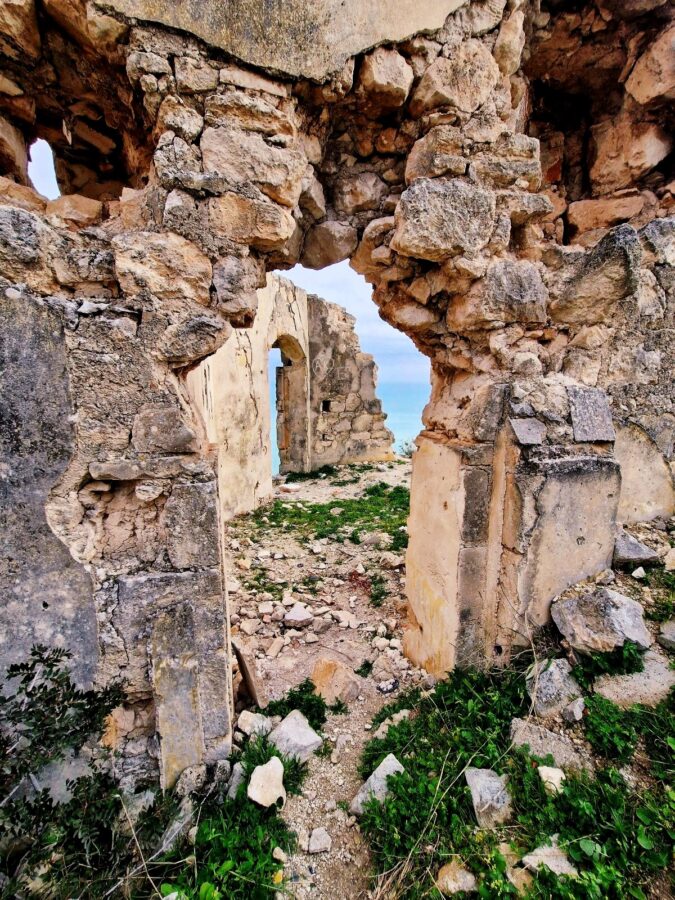 Interior view of heavily weathered, crumbling stone archways and walls filled with loose rubble and patches of green grass, looking out toward a blue sea in the distance.
