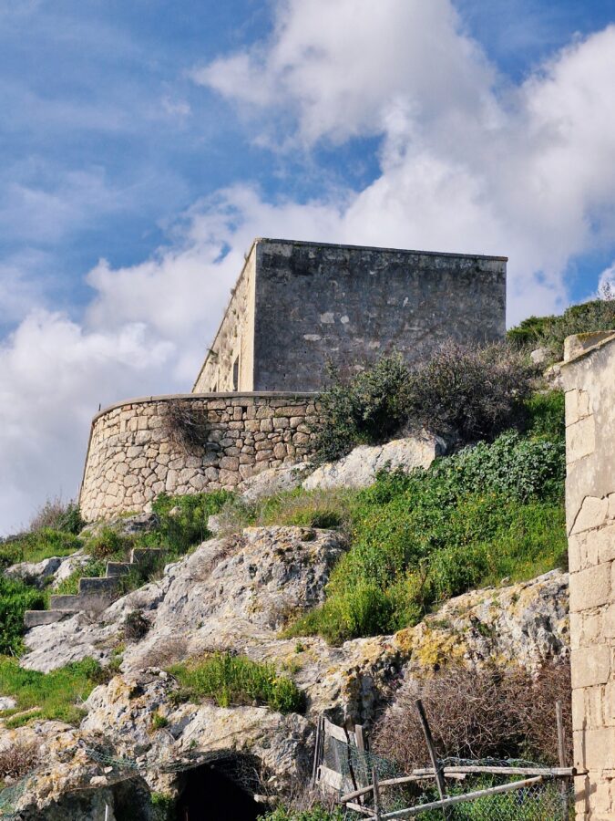 A low-angle view of an old stone structure built atop a rocky, grassy hillside. The building features a large, rectangular weathered concrete wall supported by a curved, dry stone retaining wall below it. Rough stone steps lead up the side of the rocky terrain, and a blue sky with fluffy white clouds fills the background.