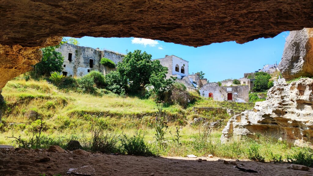 View from inside a cave looking out at stone and white buildings set against a green valley in Gravina di Ginosa, Puglia.