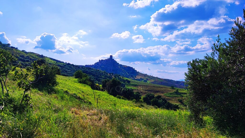 Distant view of the ghost town of Craco in Basilicata, Italy, perched on a hilltop surrounded by green rolling hills and a bright blue sky with scattered clouds