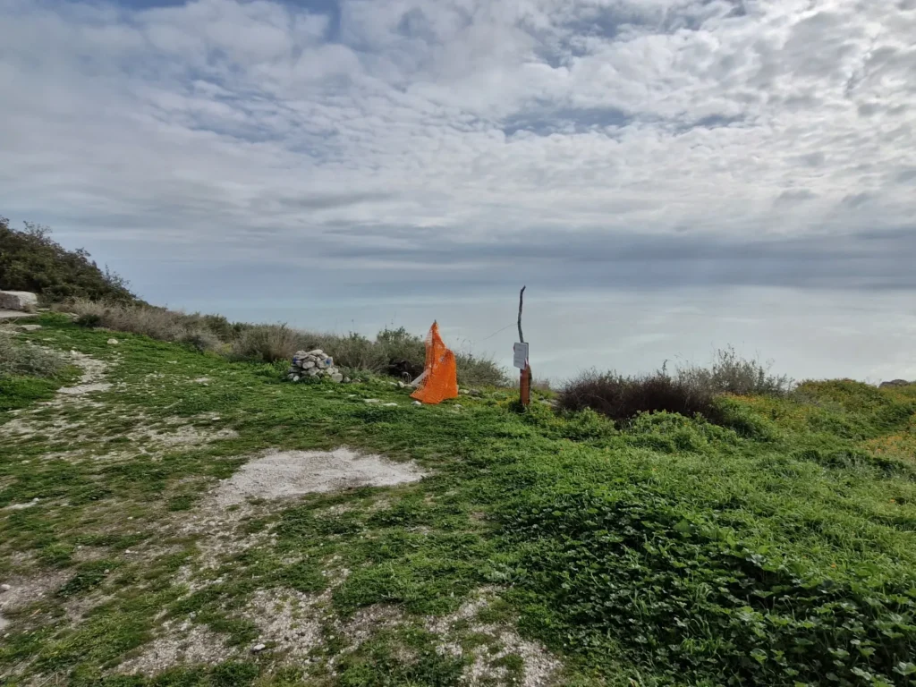 A wide landscape view of a grassy hilltop trail overlooking a hazy, pale blue sea under a sky full of white clouds. In the middle ground, a makeshift wooden post with a small white sign stands near a piece of bright orange safety netting, marking a spot along the vegetated edge of the cliff.