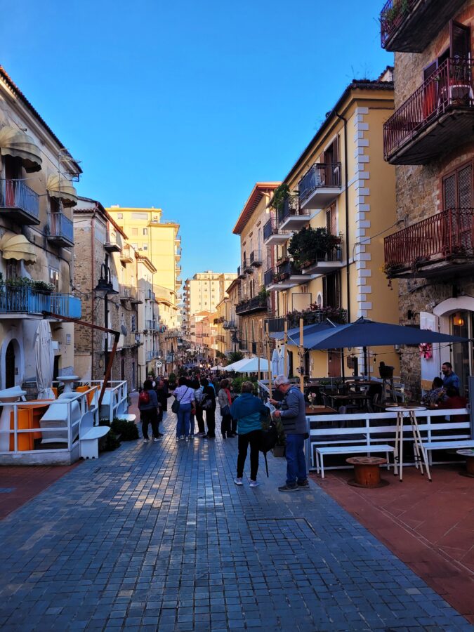 Narrow pedestrian street with stone buildings, outdoor restaurant tables, and people strolling in Agropoli, Campania