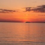 Orange sunset over the Tyrrhenian Sea seen from Agropoli on the Cilento Coast, with a dark island silhouette on the horizon and calm water reflecting the evening sky.