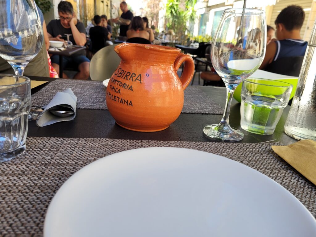 An outdoor restaurant table setting featuring a terracotta ceramic pitcher with the text "Sbirra Trattoria Tipica Napoletana," surrounded by wine glasses, water glasses, and a white plate on woven placemats.