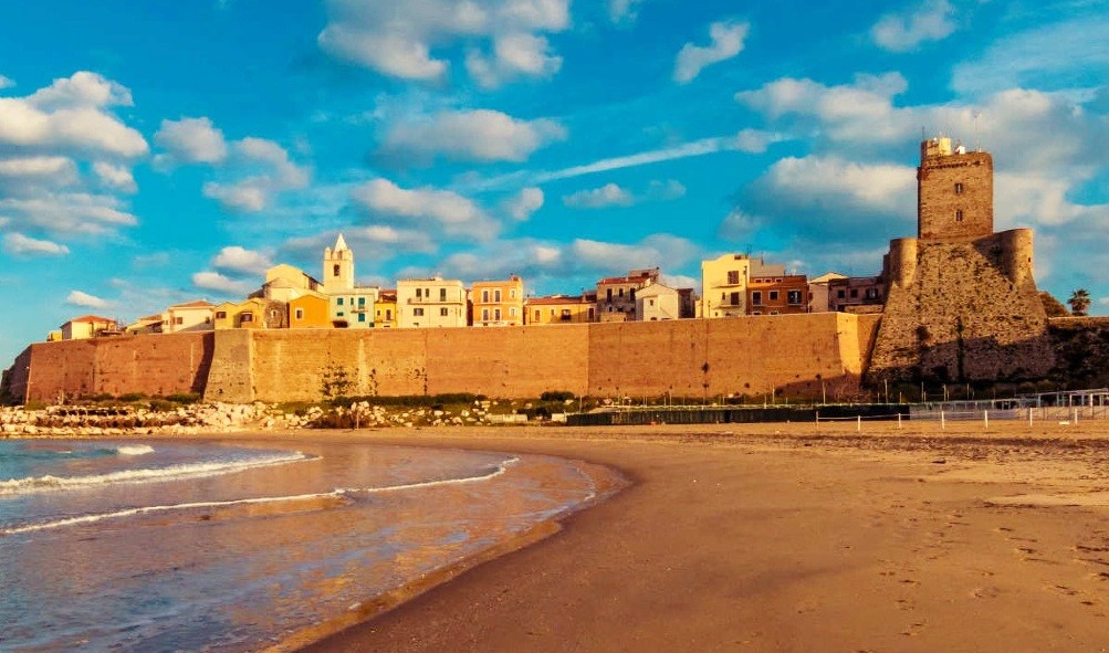 A wide-angle view of the Termoli old town (Borgo Antico) fortified walls and the Swabian Castle (Castello Svevo) seen from the sandy beach at sunset under a blue sky with scattered clouds.