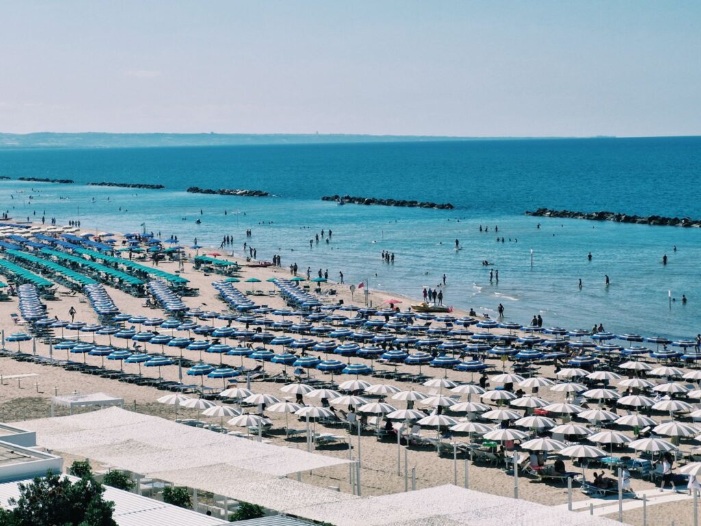 A high-angle view of a crowded sandy beach in Termoli, Molise, lined with long, symmetric rows of blue, turquoise, and white beach umbrellas and sun loungers next to a calm blue sea.