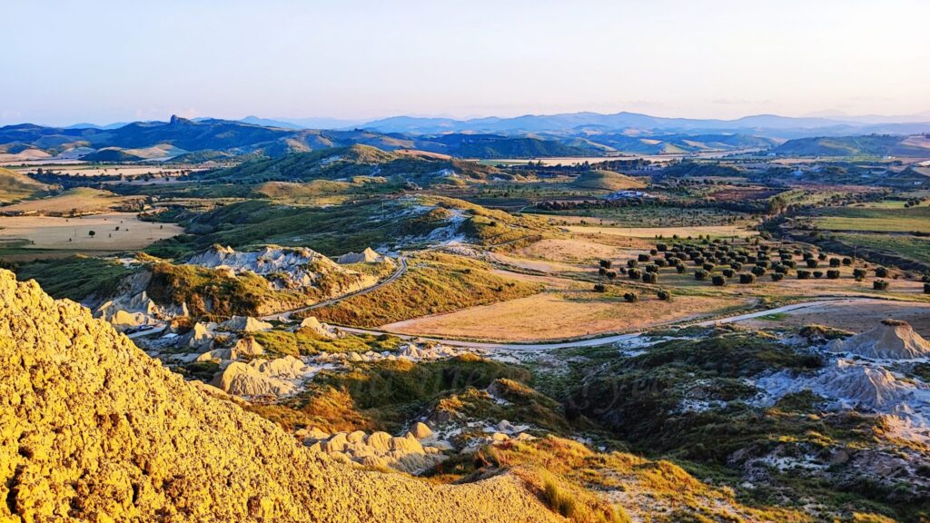 Panoramic view from the top of the Calanchi in Pisticci, Basilicata, Italy. In the foreground, steep, clayey, yellowish-gray rock formations with an irregular, rough texture are illuminated by warm afternoon sunlight. In the background, a rolling valley landscape unfolds with cultivated fields, patches of olive trees, and a winding narrow road. Mild ridges and distant mountains line the horizon under a clear, bright sky.