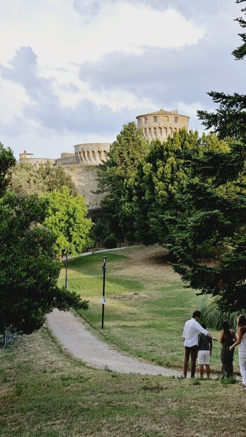 A view of the large, circular stone towers and fortified walls of the Fortezza Medicea in Volterra, Tuscany, rising behind a green park. In the foreground, a winding gravel pathway cuts through a grassy hill dotted with trees, with a small group of people standing on the right under a cloudy sky.