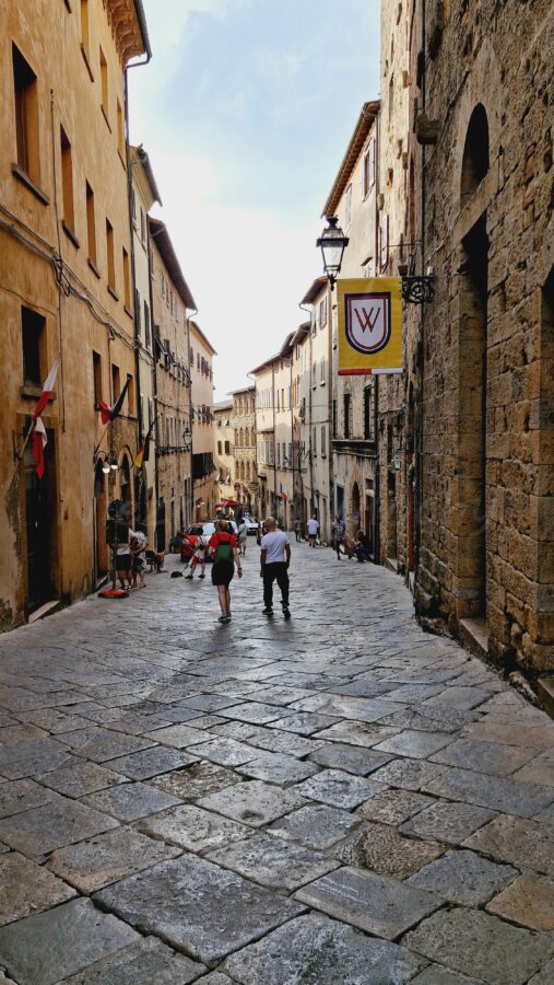 A narrow, sloping street paved with large, uneven stone slabs in Volterra, Tuscany, flanked by tall, old stone and stucco buildings. Pedestrians walk along the street, which features historic flags, a vintage metal streetlamp, and a yellow banner with a red and white crest hanging from a wall.