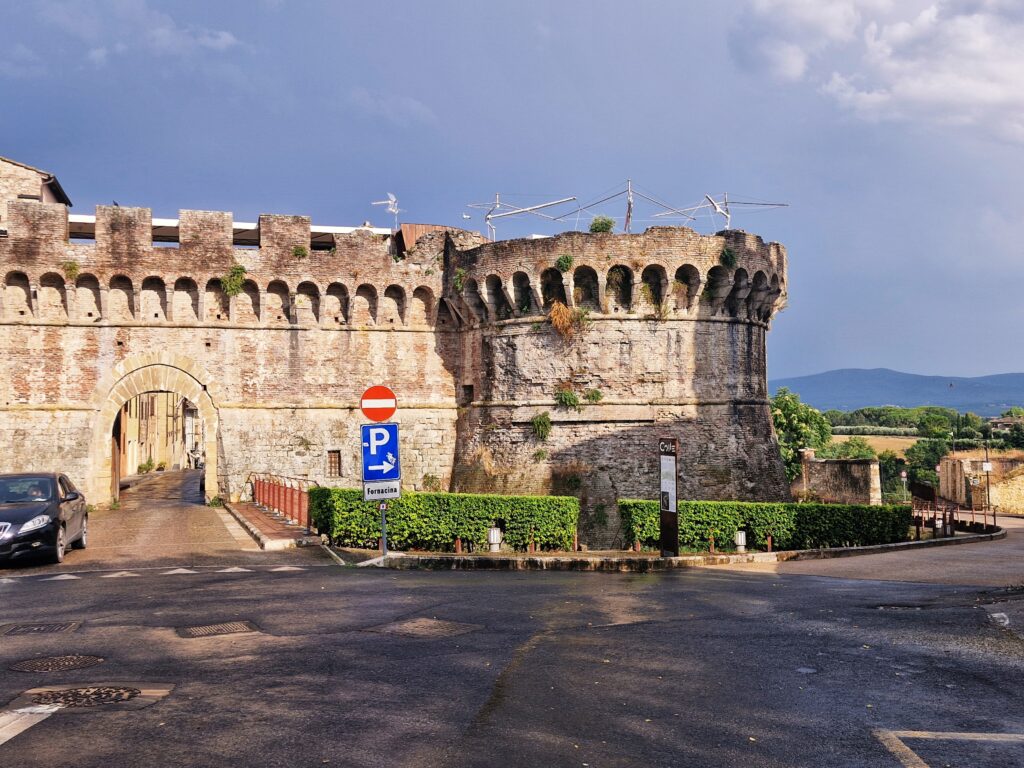 The medieval stone fortification walls of Colle di Val d'Elsa, featuring a large rounded bastion on the right and an arched gateway on the left. In the foreground, there is a wet asphalt street, a blue parking sign next to a low green hedge, and a dark car partially visible on the far left.