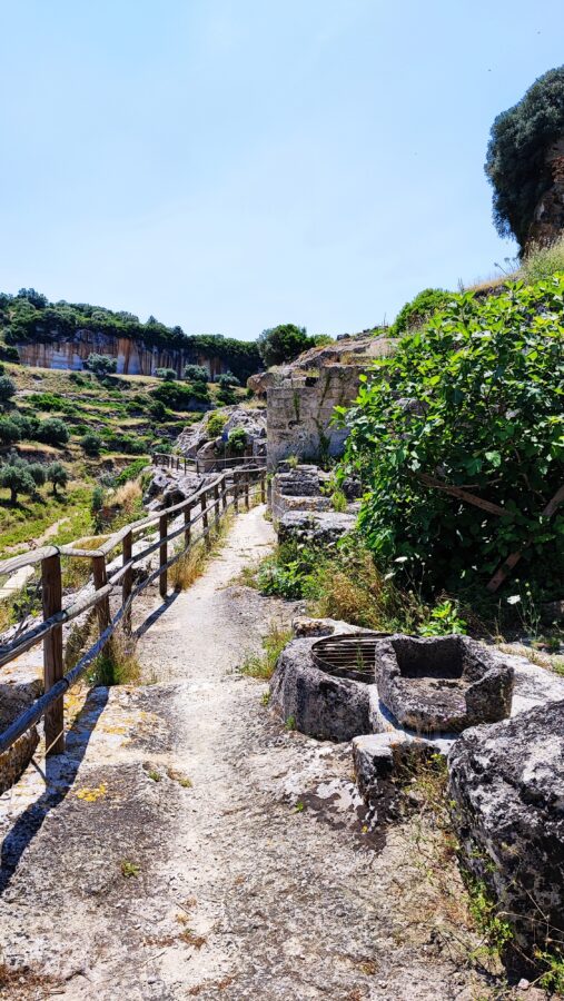 Stone path with a wooden railing leading along the edge of the canyon toward the troglodyte cave settlement in Gravina di Ginosa, Puglia.