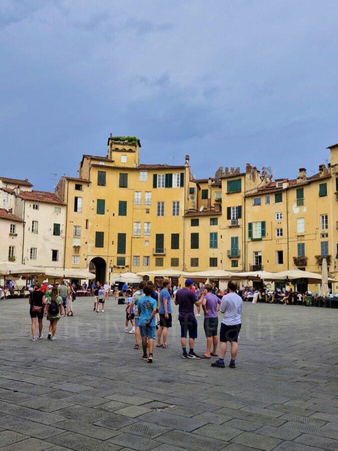 A view of the wide, oval-shaped Piazza dell'Anfiteatro in Lucca, Italy, surrounded by tall, yellow historic buildings with green window shutters. In the center of the stone-paved square, groups of tourists gather, while outdoor restaurant tables with large white umbrellas line the base of the buildings under a dramatic, overcast blue-grey sky.