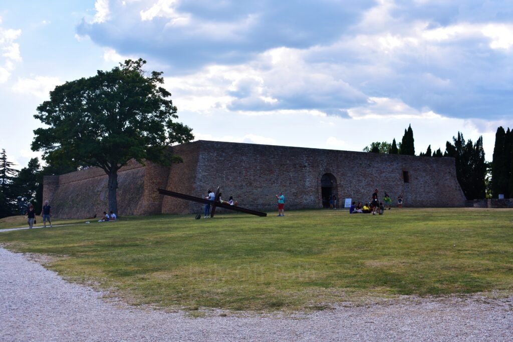 Fortezza Albornoz brick fortress and Parco della Resistenza lawn in Urbino, with visitors sitting near a modern metal sculpture.