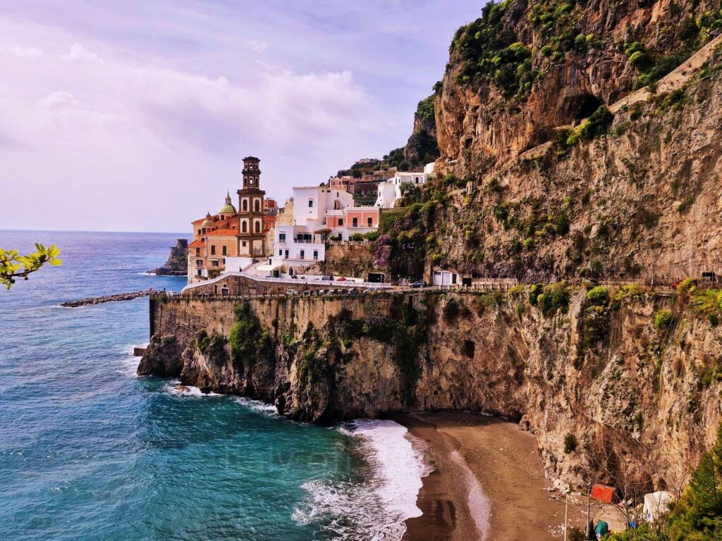 Path of the Lemons views. Coastal village of Atrani perched on steep cliffs along the Amalfi Coast, featuring white buildings, a church tower, and a small sandy beach below the road.