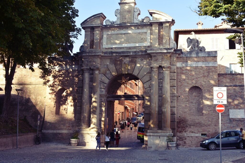 Porta Valbona, a large stone city gate with classical columns and an arched entrance leading into the historic center of Urbino, Marche, Italy.