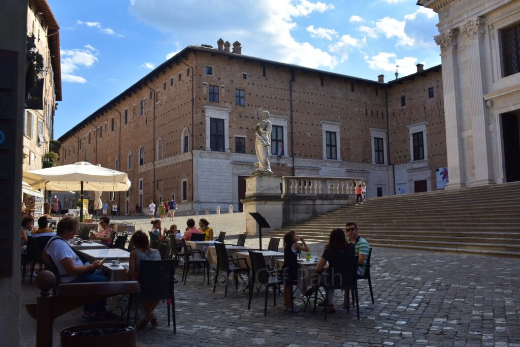 Outdoor café seating on the cobblestone square of Piazza Duca Federico in one day in Urbino, Marche, with the Ducal Palace and Cathedral steps in the background.