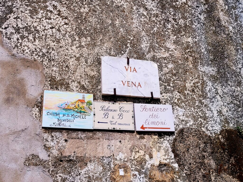 Street signs on a rough stone wall showing Via Vena and a ceramic directional sign pointing left toward the Sentiero dei Limoni trail.