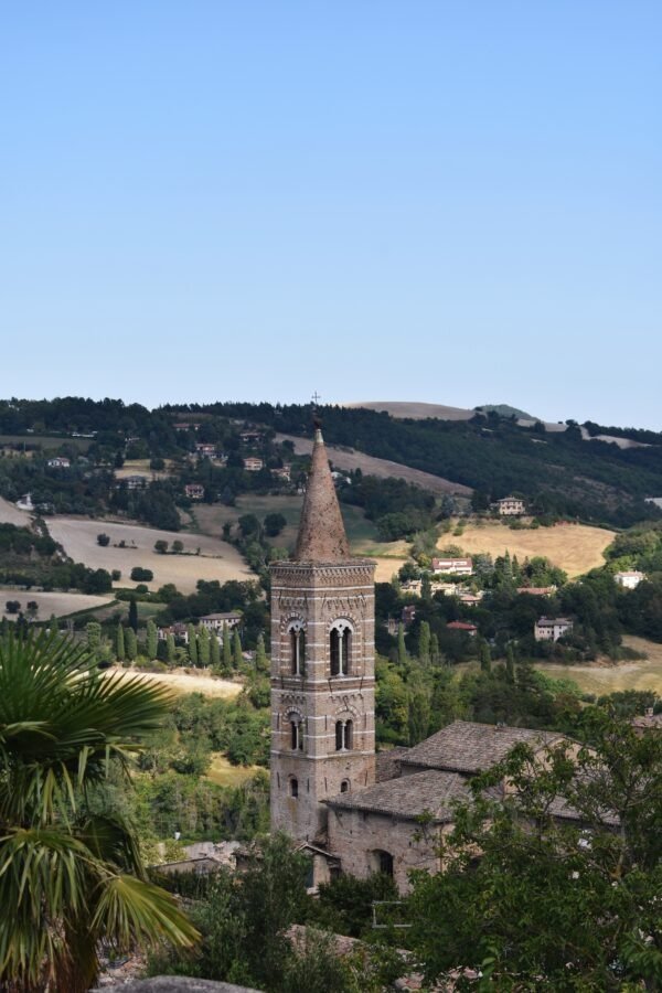 Brick bell tower of a stone church in Urbino with arched windows and a conical spire, overlooking the rolling hills of the Marche countryside.