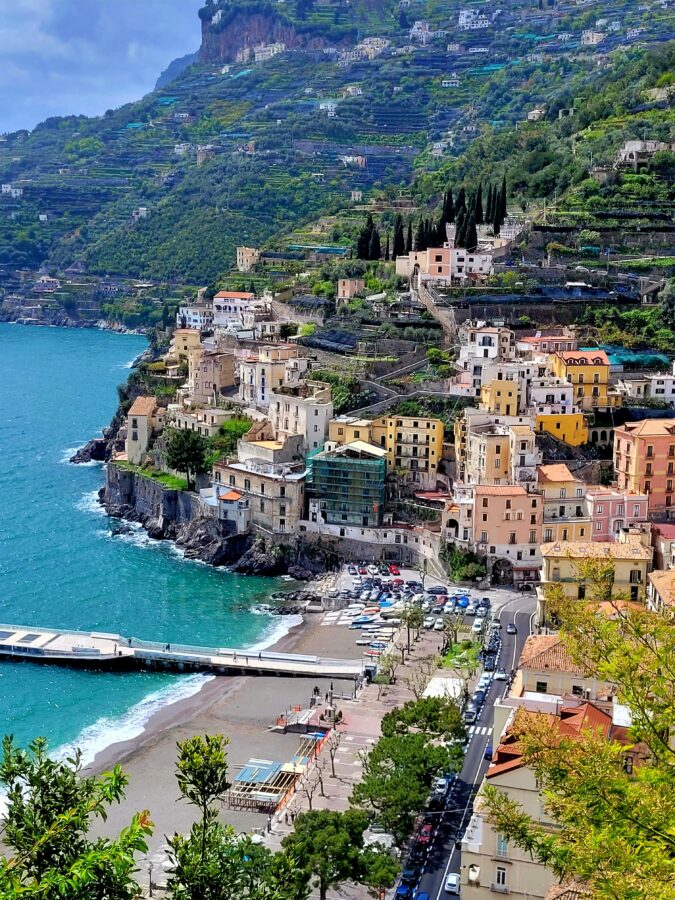 High-angle view from the Sentiero dei Limoni of Minori, showing colorful buildings along the coastline, a small beach, a pier extending into turquoise sea, and terraced lemon groves climbing the mountain behind.