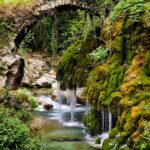 The river flows between the stones, plants grow on the banks, and an old stone bridge overgrown with greenery stands over the river. Easy hikes in Campania