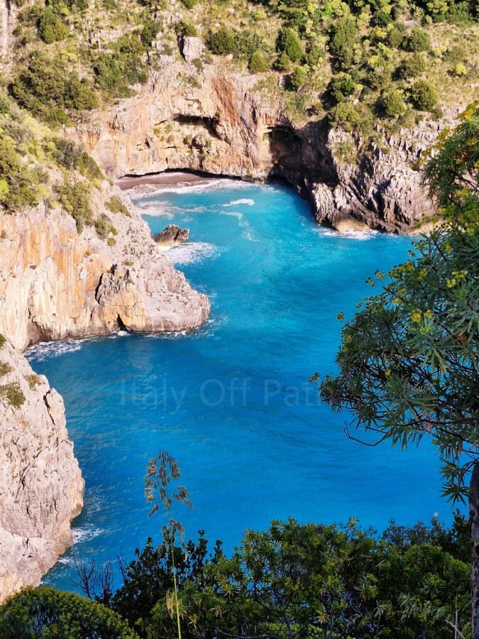 High-angle view of a rocky sea cove with turquoise water framed by steep cliffs and coastal vegetation along Sentiero Apprezzami l'Asino in Cilento, Campania, Italy.