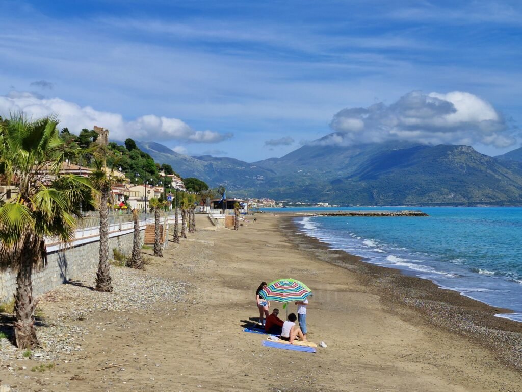 Small group sitting under a striped umbrella on a wide, empty dark sand beach in Campania, Italy, with palm trees and mountains in the background.