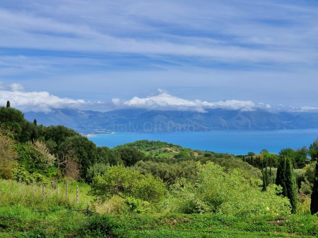 Elevated view over green hills and Mediterranean vegetation toward the sea and distant coastal mountains under a cloudy sky in Campania, Italy.