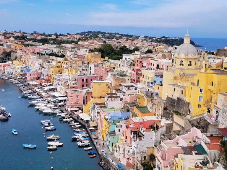 yellow, blue, white and orange houses stand in a port by the sea, where you can see boats. Behind the houses you can see the sea in Procida Island Italy
