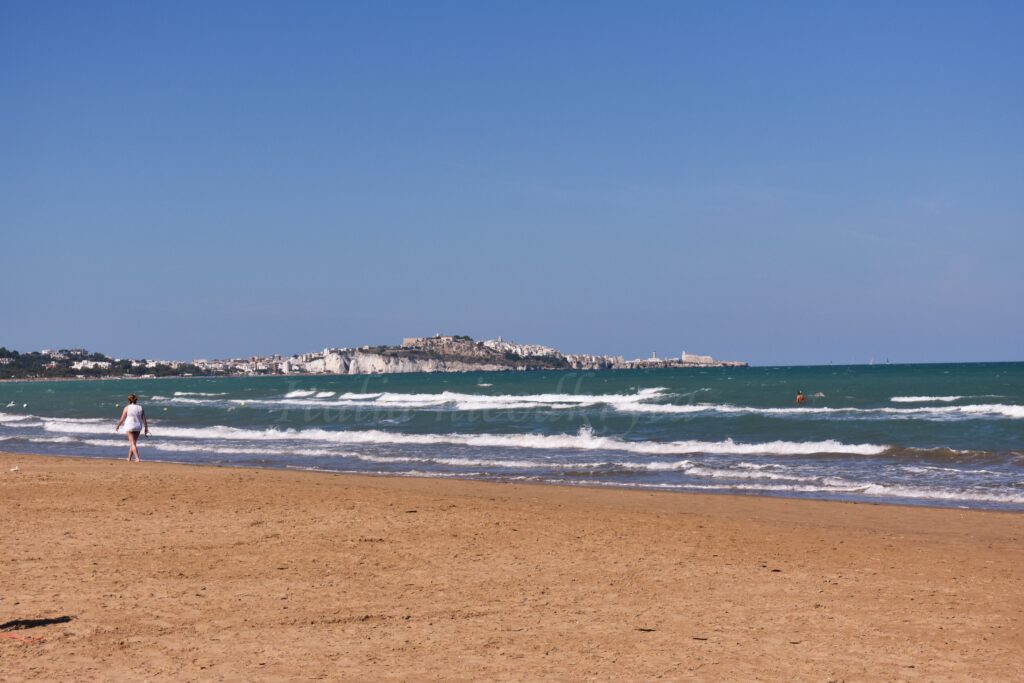 Szeroka piaszczysta plaża w Vieste w Apulii, z falującym morzem Adriatyckim i widokiem na stare miasto położone na białych klifach w oddali pod błękitnym niebem