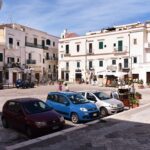 Plac otoczony białymi niskimi budynkami z zielonymi okiennicami, przed budynkami parkują samochody osobowe.