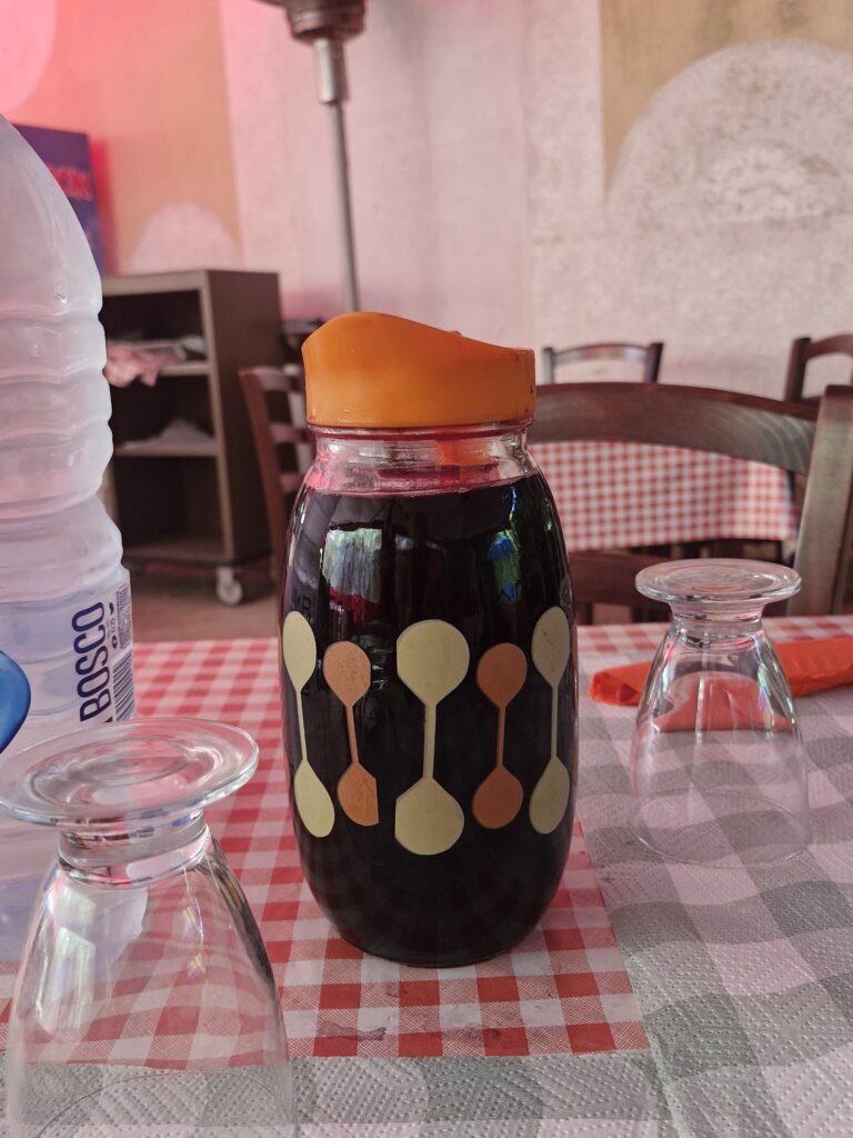 Dzbanek czerwonego wina na plastikowym stole z ceratą w restauracji Punto di Ristoro La Cascata, z odwróconymi szklankami i prostym, prowincjonalnym wystrojem w tle.