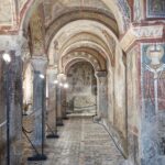 Interior of a medieval crypt with arched ceilings richly decorated with frescoes and patterned mosaic floors, softly illuminated by modern standing lights.