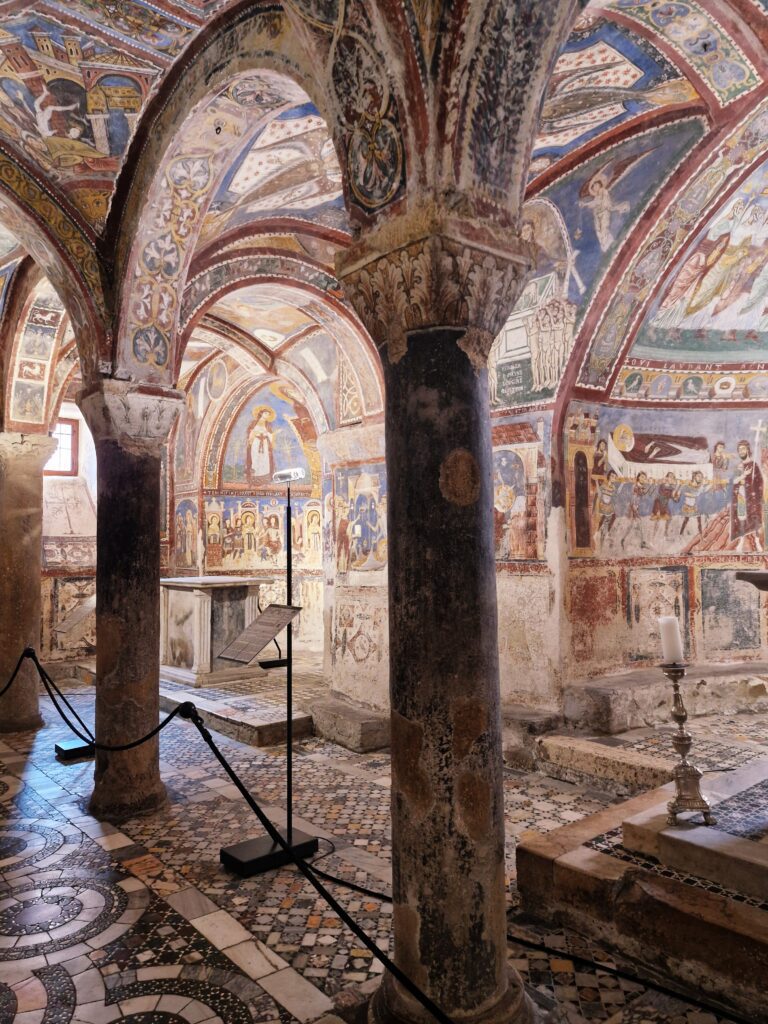 What to see near Rome.Interior of a medieval crypt with frescoed arches, stone columns, and intricate mosaic floors. Description: The image captures the richly decorated interior of an ancient crypt. The vaulted ceiling