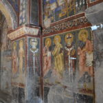 Medieval frescoes inside a stone church depicting saints and biblical figures with halos, vibrant robes, and Latin inscriptions.