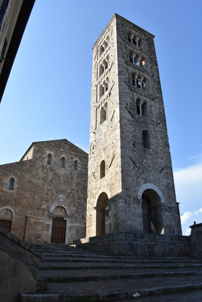 Wysoka kamienna wieża z małymi oknami i drzwiami stoi przed kamienna katedrą.