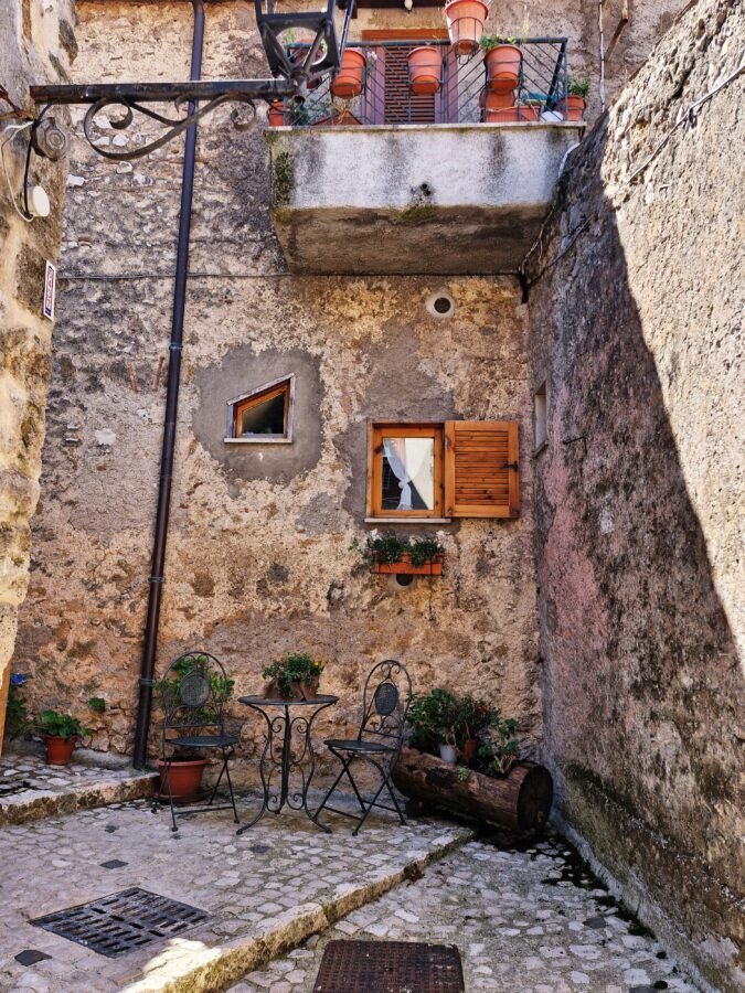 Cobblestone courtyard in Trevi nel Lazio with two wrought-iron chairs, a small table, rustic stone walls, and small shuttered windows beneath an overhead balcony.