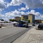 Street scene with a blue bus, motorbike, and people gathered near tents in front of modern apartment buildings under a partly cloudy sky.
