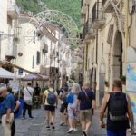 Busy pedestrian street in an Italian town with festival lights overhead, people walking among shops and cafes, and green mountains rising in the background.