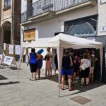 Group of people standing under a canopy tent in an Italian town square, waiting in line for an event or registration.