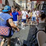 Crowd of people walking barefoot through a shallow stream of water flowing along a narrow Italian street during a summer event, with a man leading a dog on a leash. A Chiena Campagna
