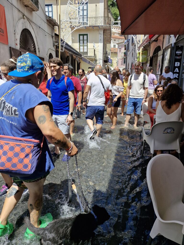 Crowd of people walking barefoot through a shallow stream of water flowing along a narrow Italian street during a summer event, with a man leading a dog on a leash. A Chiena Campagna