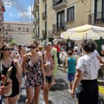 Crowds of people walking barefoot through shallow water flowing in a sunny Italian street lined with balconies, umbrellas, and festive arches.