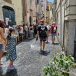 People walking barefoot through shallow water flowing along a narrow Italian street lined with pastel-colored old buildings.
