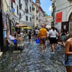 Ludzie brodzący w wodzie na zalanej ulicy podczas święta A Chiena Campagna w Kampanii; niektórzy siedzą w kawiarniach, inni niosą niebieskie wiadra, a w tle widać stare kamienice i wieżę kościoła