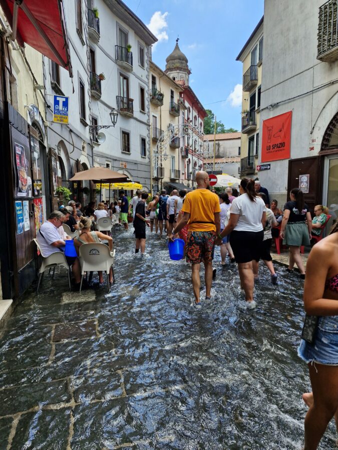 Ludzie brodzący w wodzie na zalanej ulicy podczas święta A Chiena Campagna w Kampanii; niektórzy siedzą w kawiarniach, inni niosą niebieskie wiadra, a w tle widać stare kamienice i wieżę kościoła