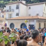 Tłum ludzi biorących udział w wodnej bitwie podczas święta A Chiena w Campagna w Kampanii; uczestnicy z niebieskimi wiadrami oblewają się wodą na tle jasnych budynków i zielonego wzgórza.